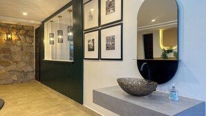 A modern bathroom with a stone wall, a sink with a decorative bowl, a mirror, and framed pictures on the wall. - Vee Serviced Apartments & Hotel (Entebbe)