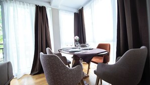 En matplats med ett bord dukat för två personer, omgiven av mönstrade stolar, stora fönster med gardiner och utsikt över grönskan utanför.