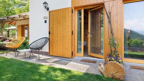 Een houten huis met een schuifdeur, een klein terras met een tafel en stoelen, en een houtblok met bloemen.