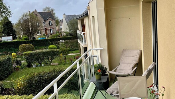 A balcony with a table set for dining, chairs, a small table with a potted plant, and a view of a garden and neighboring houses. - Apartment in quiet, secure residence (Paimpol)