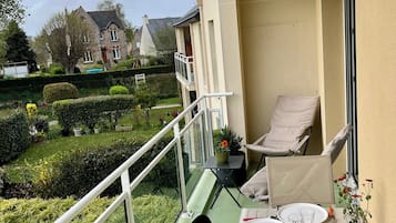 Un balcon avec une table dressée pour le repas, des chaises, une petite table sur laquelle est posée une plante en pot, et une vue sur un jardin et les maisons voisines.