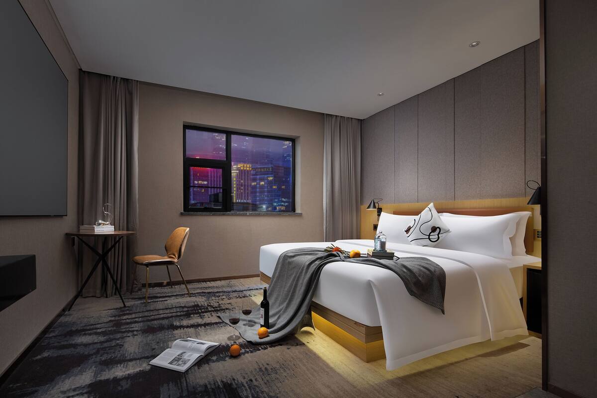 Habitación de hotel moderna con una cama grande, un escritorio y vistas a un paisaje urbano de noche.