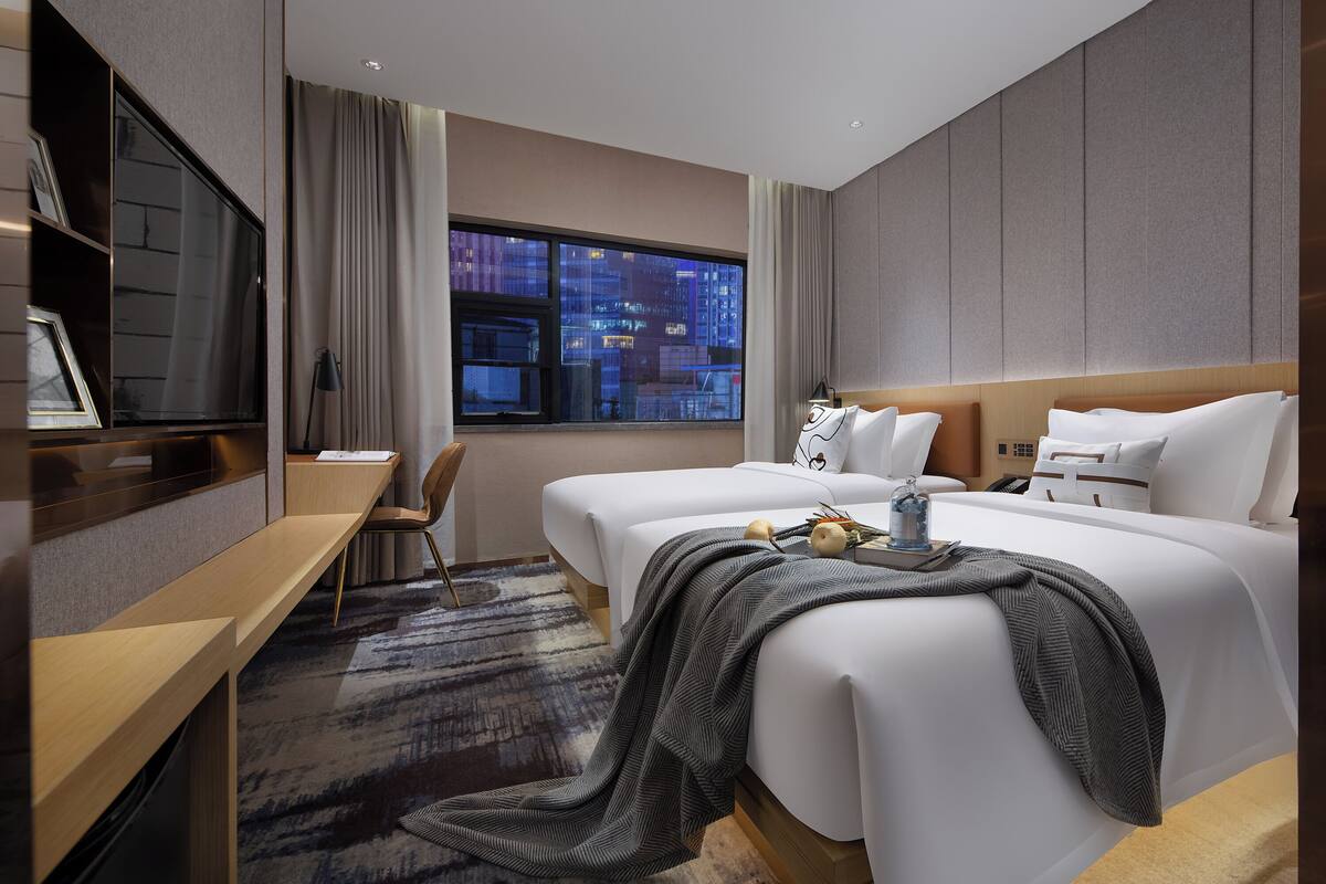 Habitación de hotel con dos camas, un escritorio, televisor y un ventanal con vistas a la ciudad.