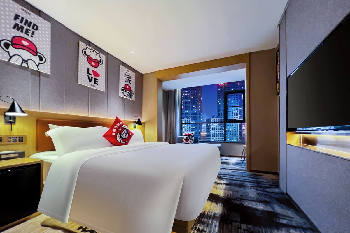 Una habitación de hotel moderna con una cama grande, televisión, vista a la ciudad y una pared con carteles de cultura popular.