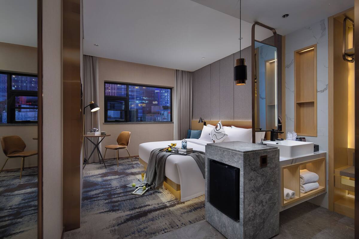 Una habitación de hotel moderna con una cama grande, un escritorio, una silla, un baño con lavamanos de mármol y vistas a la ciudad.