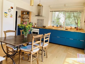 Une cuisine moderne avec des meubles bleus, une table à manger en bois avec des chaises, un vase de fleurs et une fenêtre donnant sur des arbres.