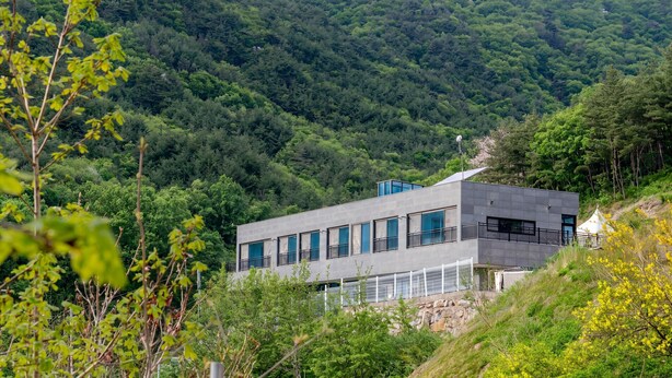 A modern building with large windows, set against a backdrop of dense forest and hills. - Sara&forest Pension Mungyeong (Mungyeong)