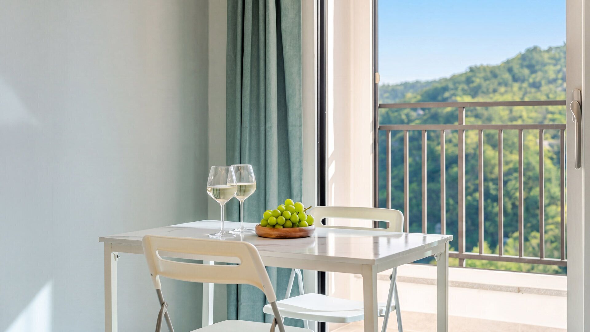 Un balcón con una mesa, dos sillas, un cuenco de uvas verdes y dos copas de vino blanco.