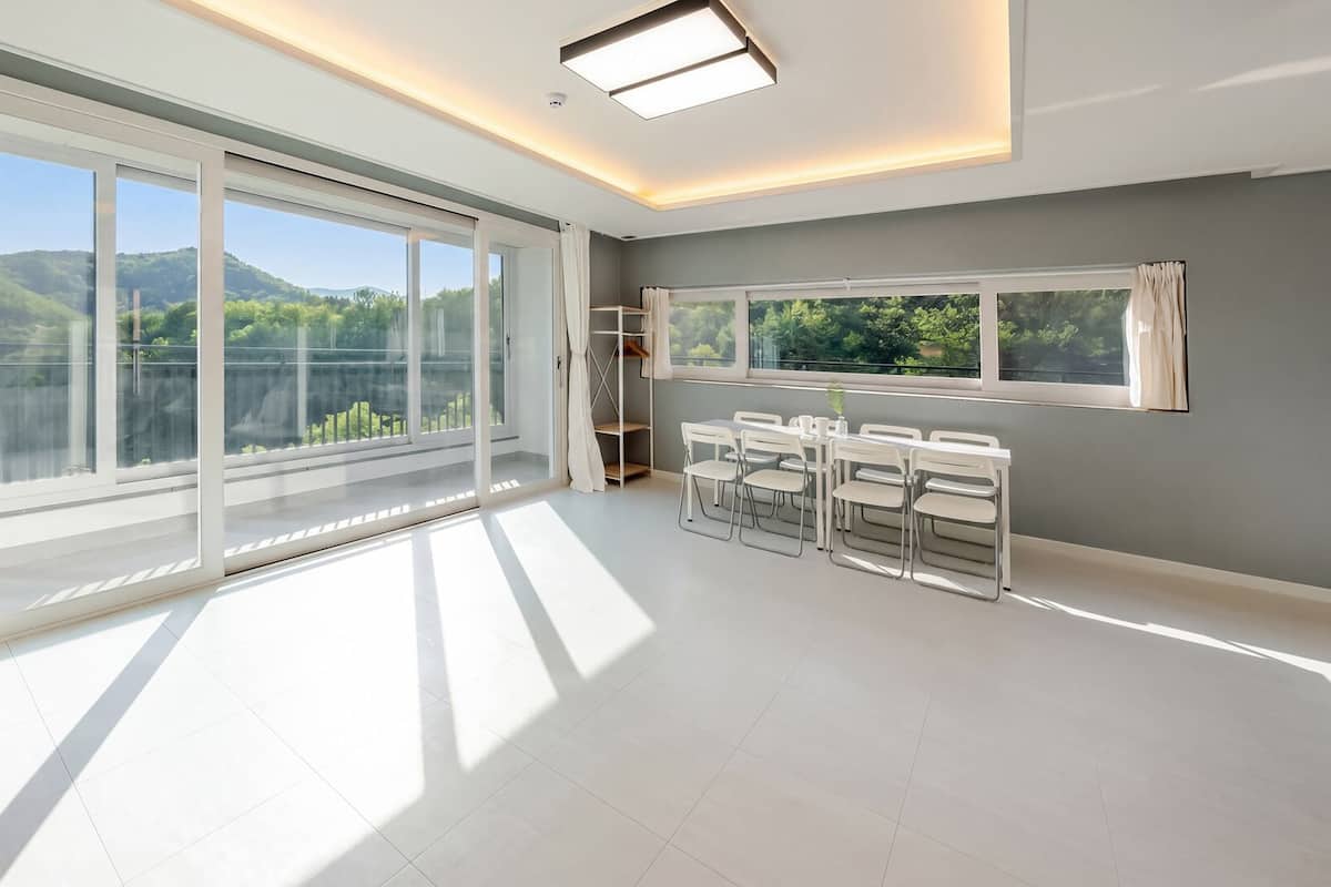 A spacious room with large windows, a dining area, and a view of greenery.