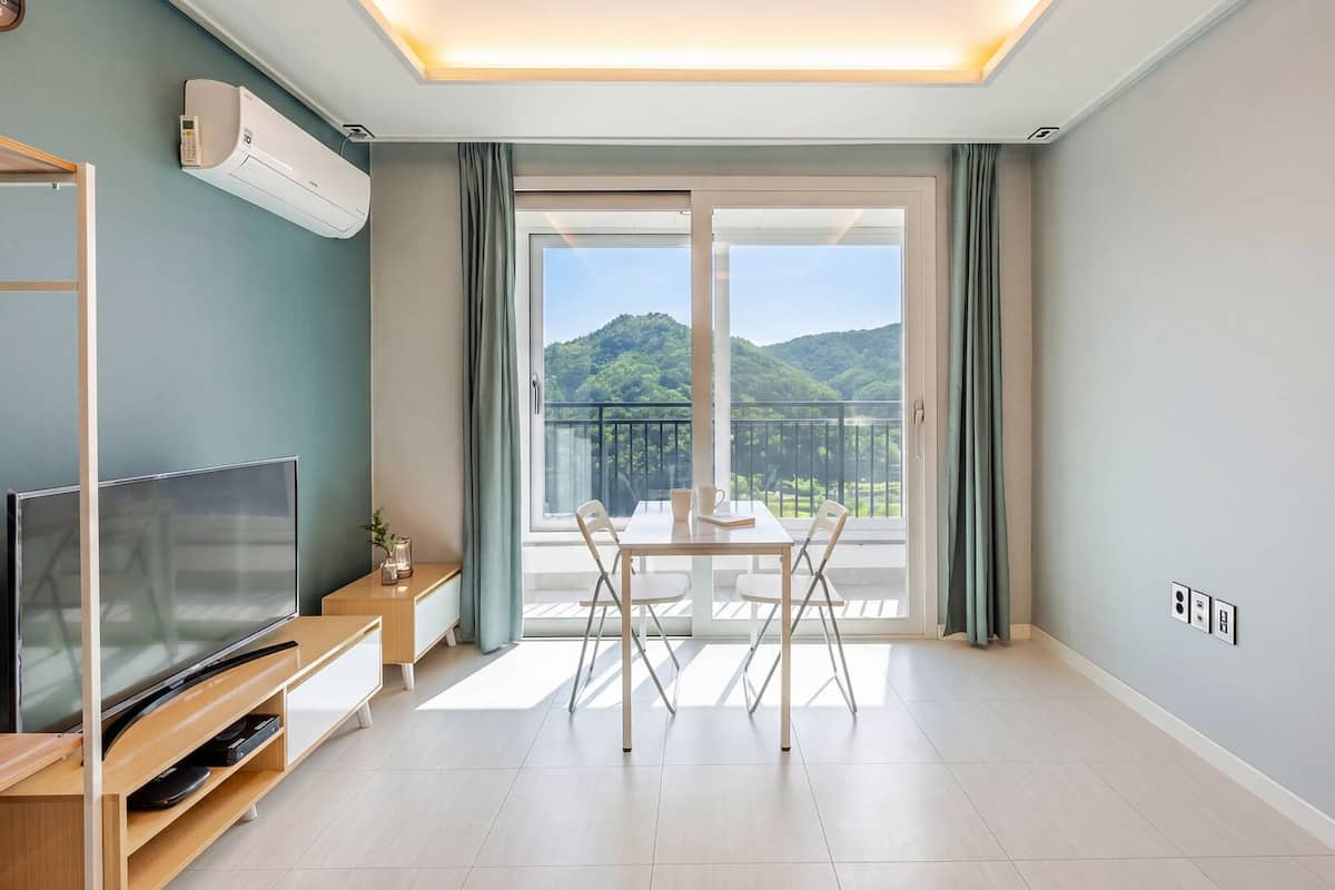 A modern living room with a flat-screen TV, a wooden entertainment unit, a dining table with chairs, and a balcony with a mountain view.