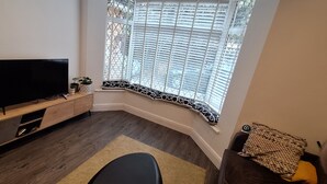 Ein Wohnzimmer mit Fernseher auf einer Holzkommode, einem Fenster mit Jalousien, einem schwarzen Sessel und einem kleinen Tisch mit einer Pflanze.