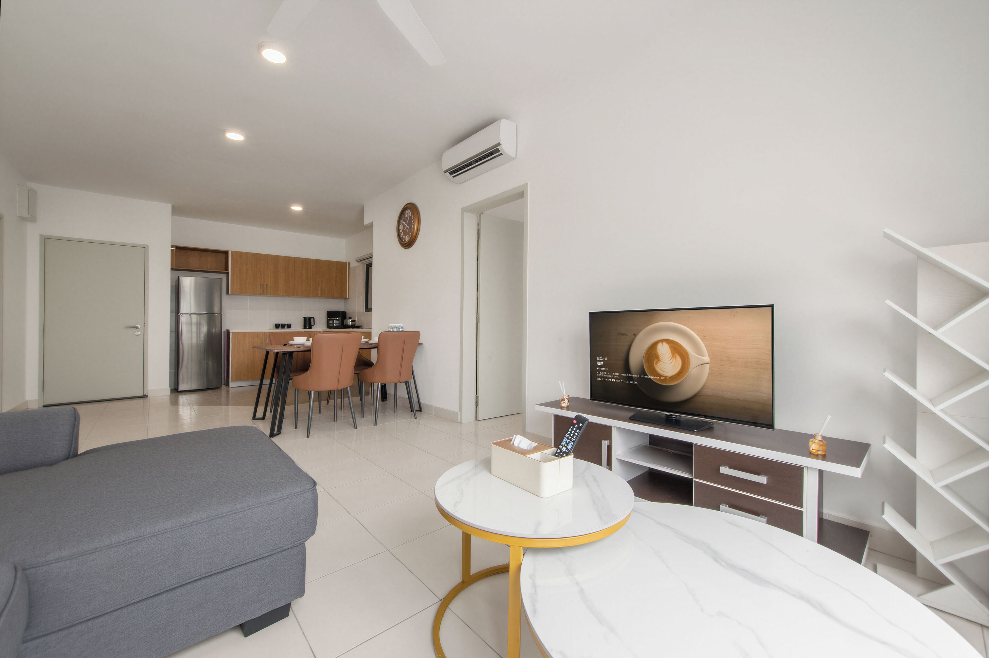 A modern living room with a sofa, a dining area with chairs, a kitchen with a refrigerator, and a TV on the wall.