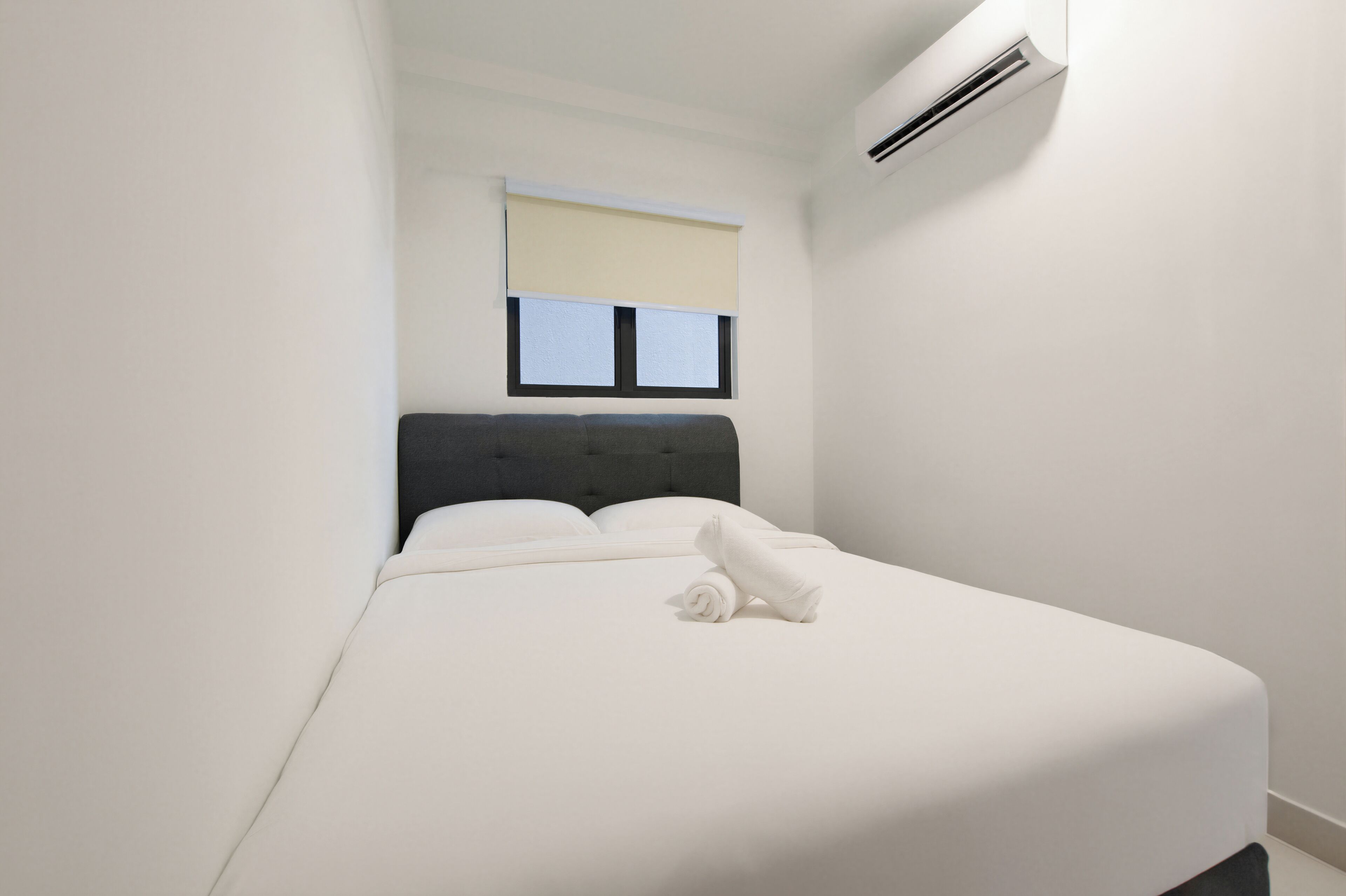 A neatly made bed with white linens and a rolled towel, a dark headboard, a window with blinds, and an air conditioning unit on the wall.