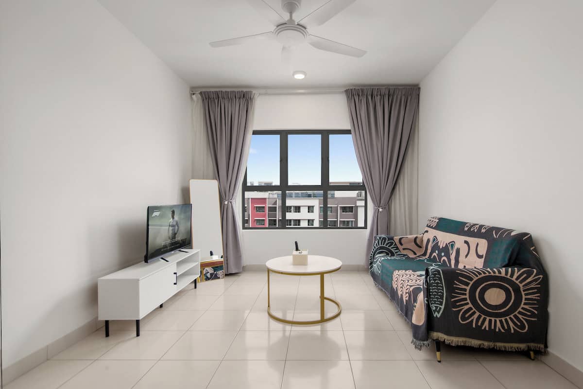 A modern living room with a flat-screen TV, a white coffee table, a sofa with a patterned throw, and a view of buildings through the window.