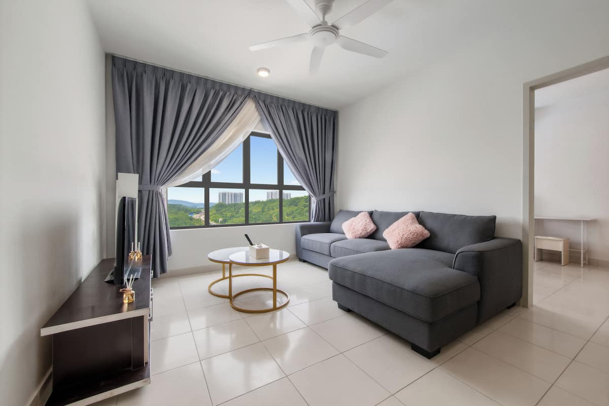 A modern living room with a grey sofa, a small round table, a ceiling fan, and a view of greenery through the window.