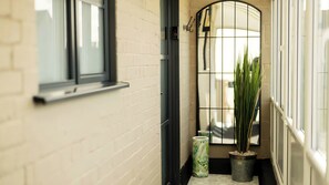 Ein schmaler Flur mit weißer Ziegelwand, einem Fenster mit schwarzem Rahmen, einem Blumentopf und einem Metallgitter.