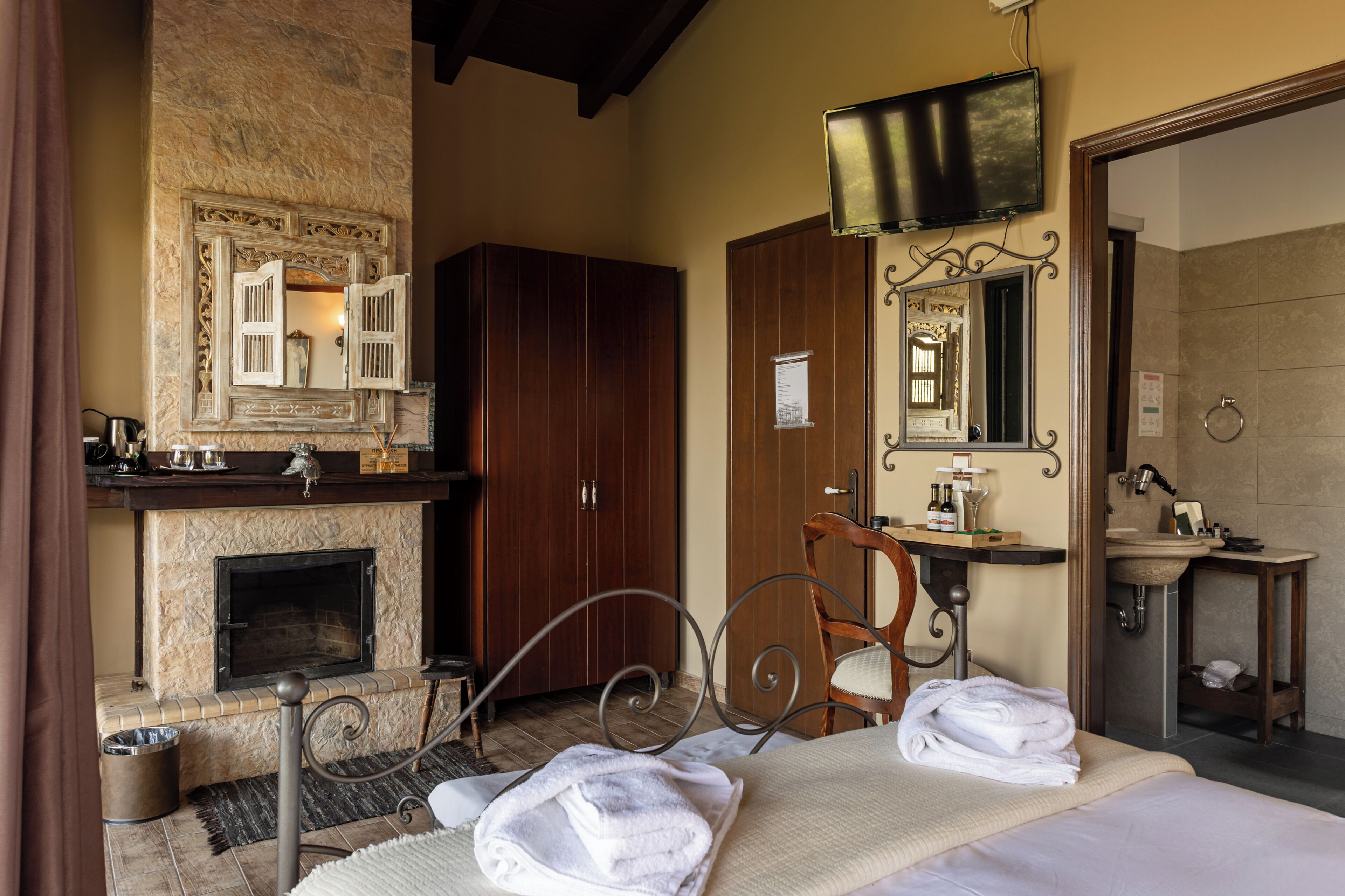 Camera d'albergo con camino, un letto corredato da asciugamani, un armadio in legno, un bagno con lavandino e specchio, e una televisione montata a parete.