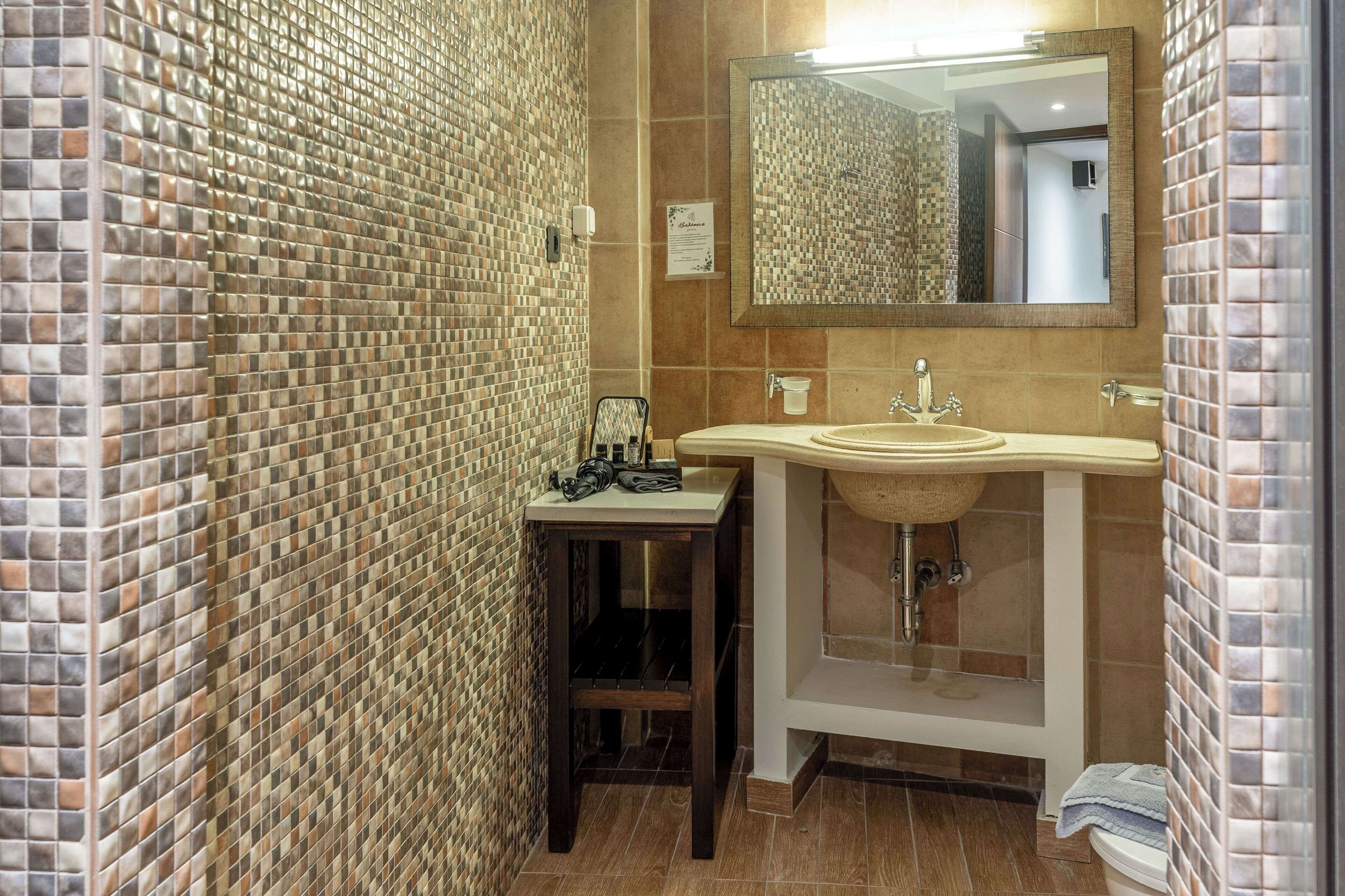 Un baño con paredes de mosaico, un lavamanos con espejo y una mesita con artículos de aseo.