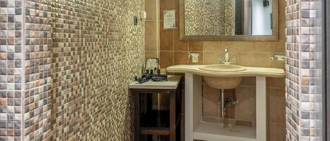 Un baño con paredes de mosaico, un lavamanos con espejo y una mesita con artículos de aseo.