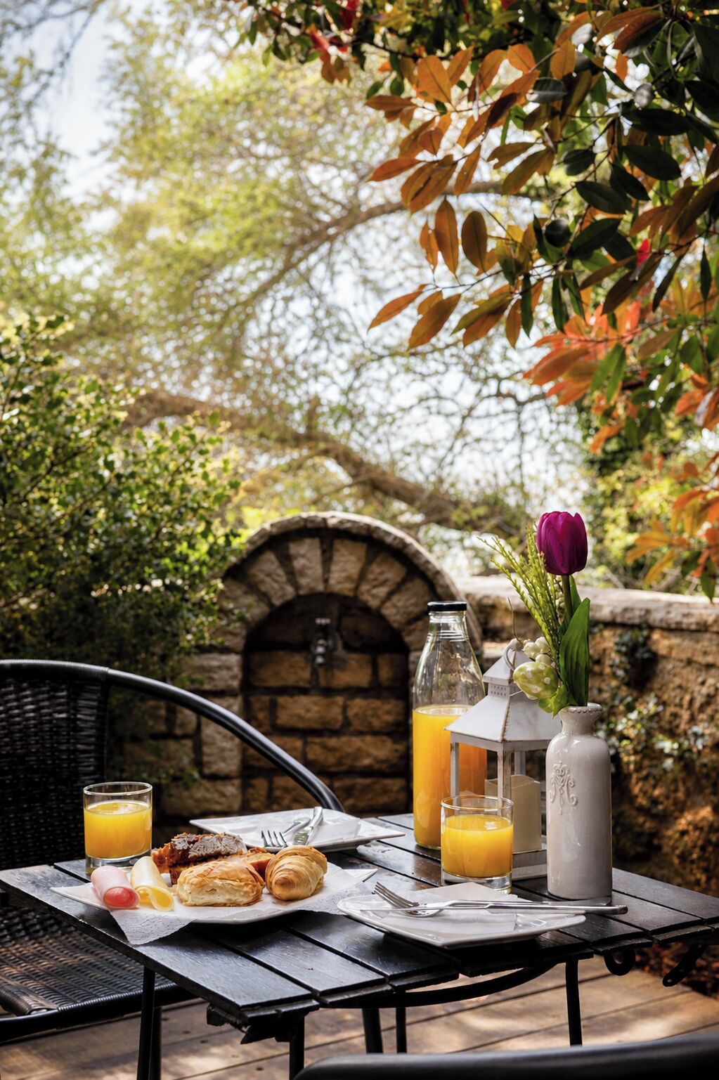 An outdoor dining area with a stone arch, a wicker chair, a table with breakfast items, and a vase with a purple flower.