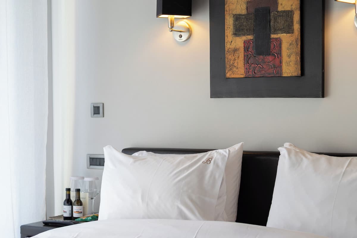 Ein Hotelzimmer mit einem ordentlich bezogenen Bett, zwei Wandlampen, einem Gemälde an der Wand und einem Nachttisch mit einer Flasche und Gläsern.