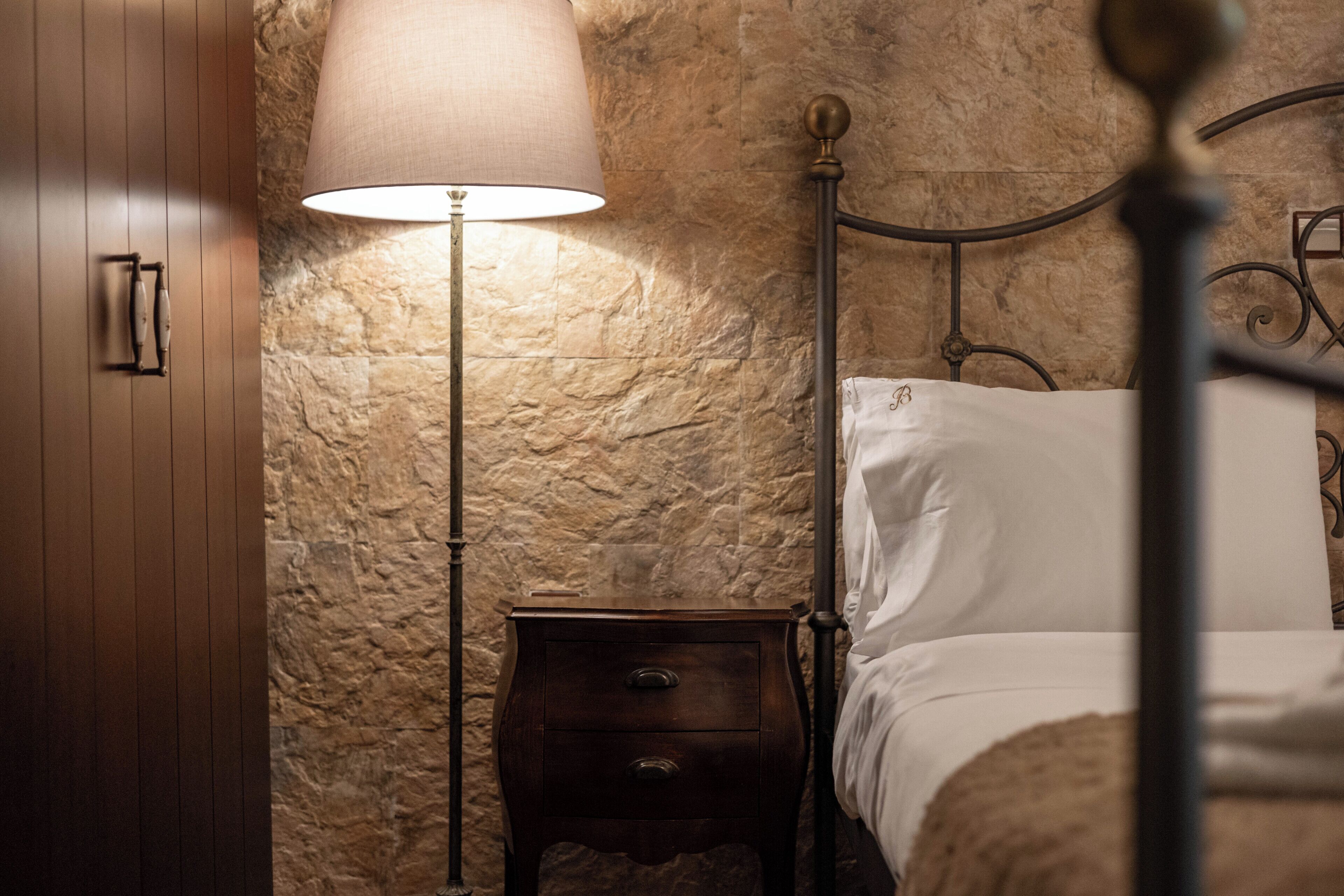 Un dormitorio con pared de piedra, mesita de noche de madera con lámpara y estructura de cama metálica.