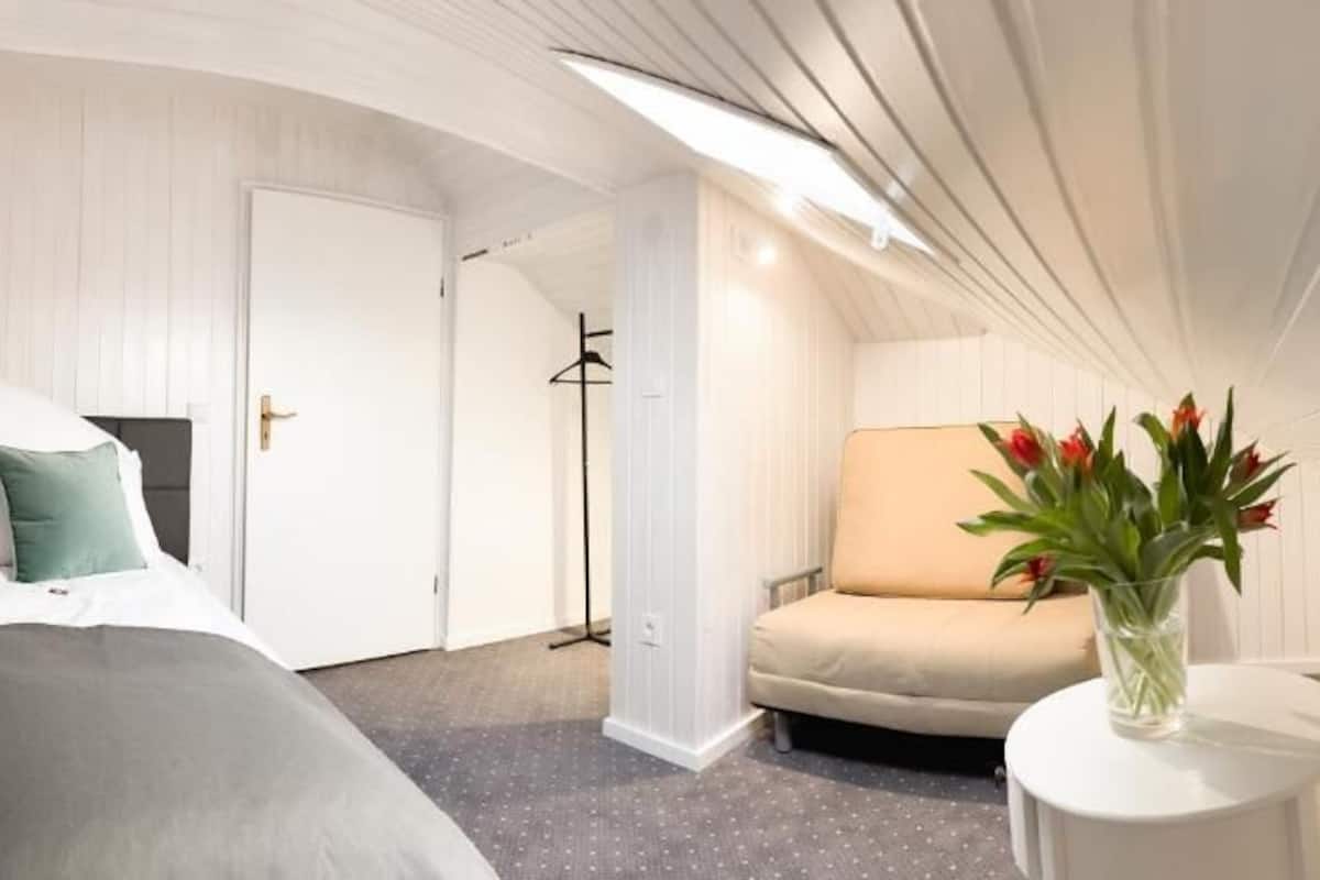 A hotel room with a bed, a chair, a small table with a vase, and a door.