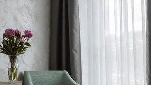 Ein Zimmer mit einem grünen Sessel, einer Blumenvase und einem Fenster mit durchsichtigen Vorhängen.