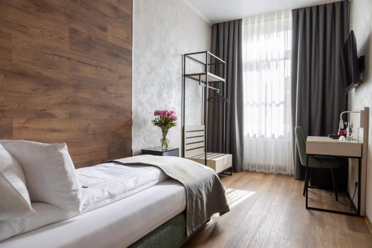 A hotel room with a bed, a desk, a chair, a TV, a window with curtains, and a plant.