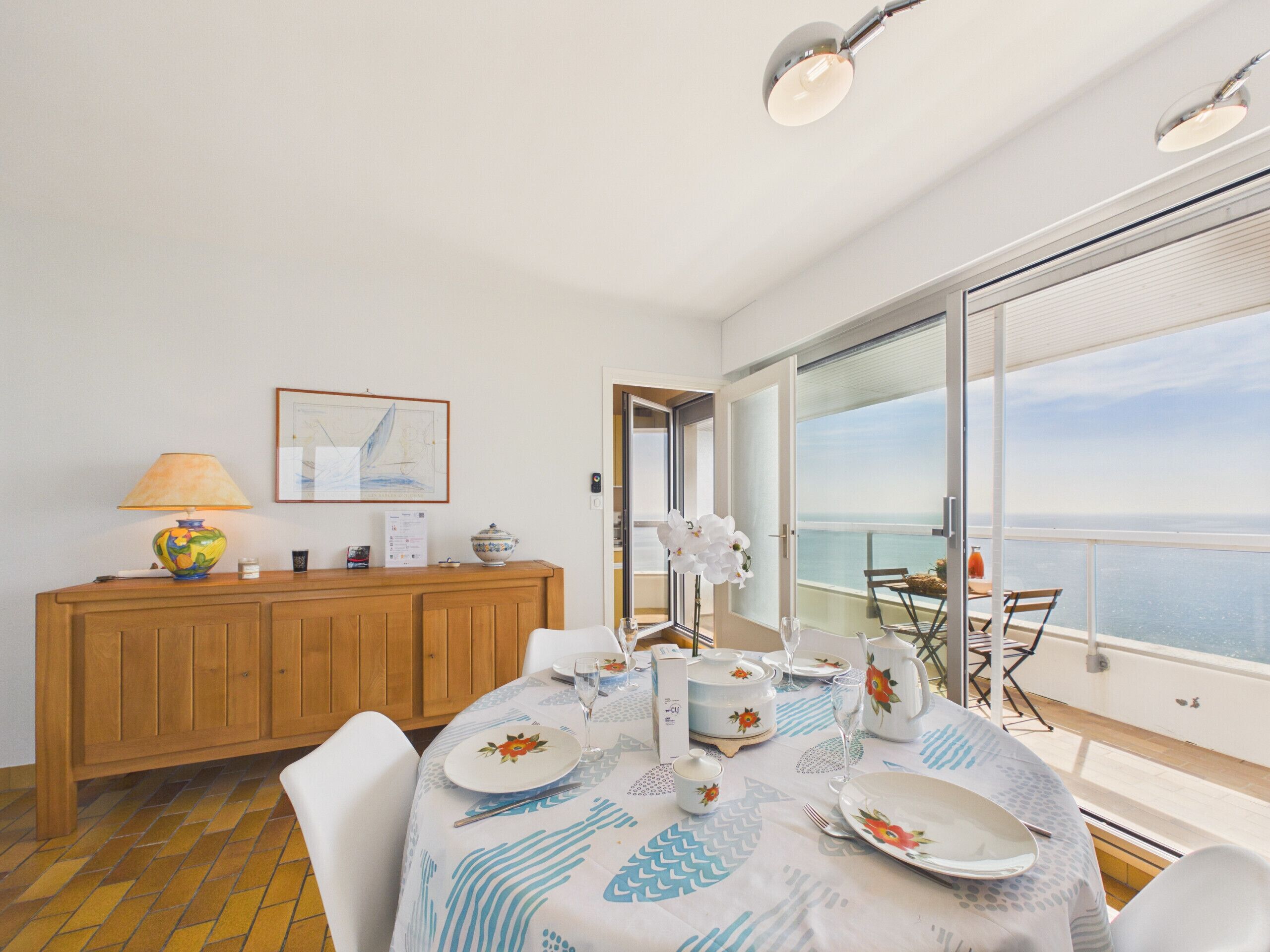 Uma sala de jantar com uma mesa posta para dois, um armário de madeira, um quadro emoldurado e vista para o mar.