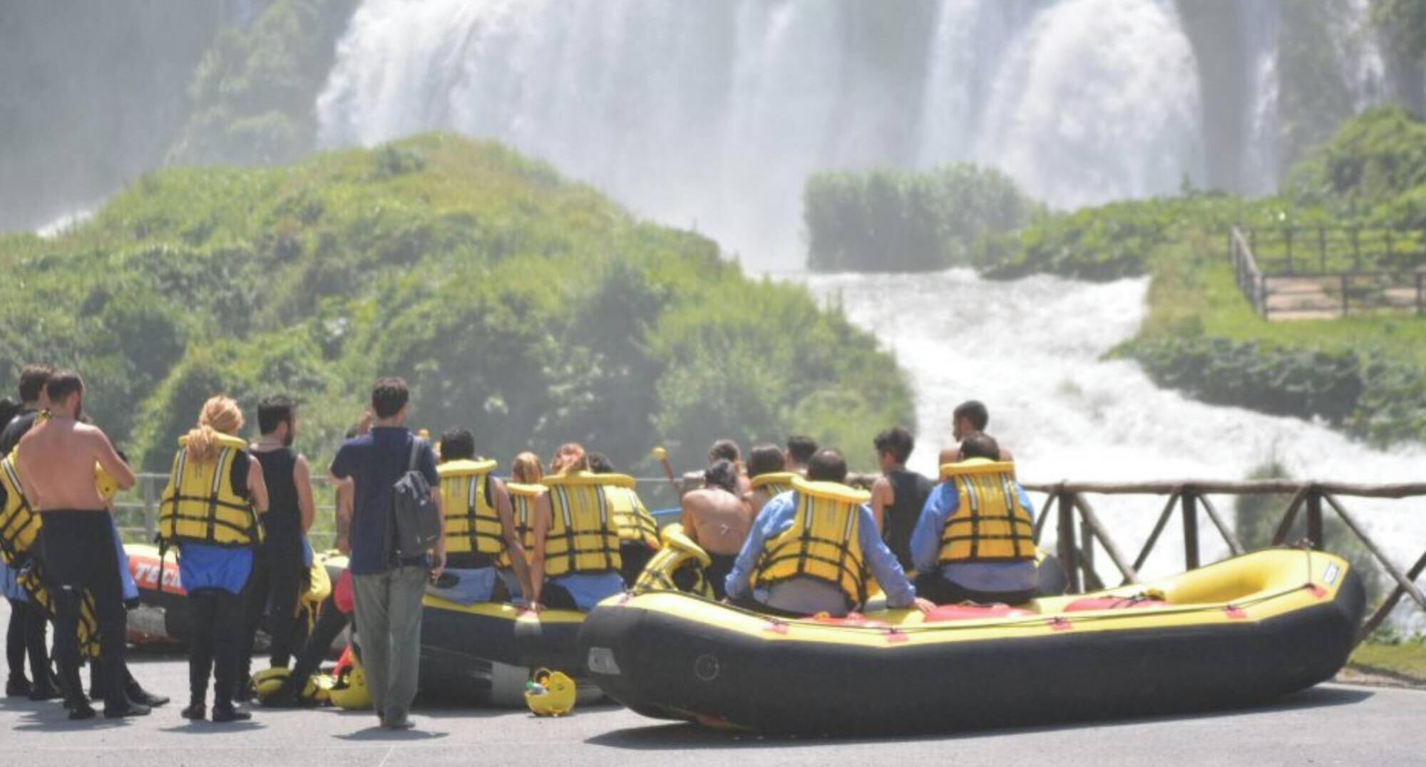 A group of people in yellow life jackets on a raft, with a waterfall in the background.