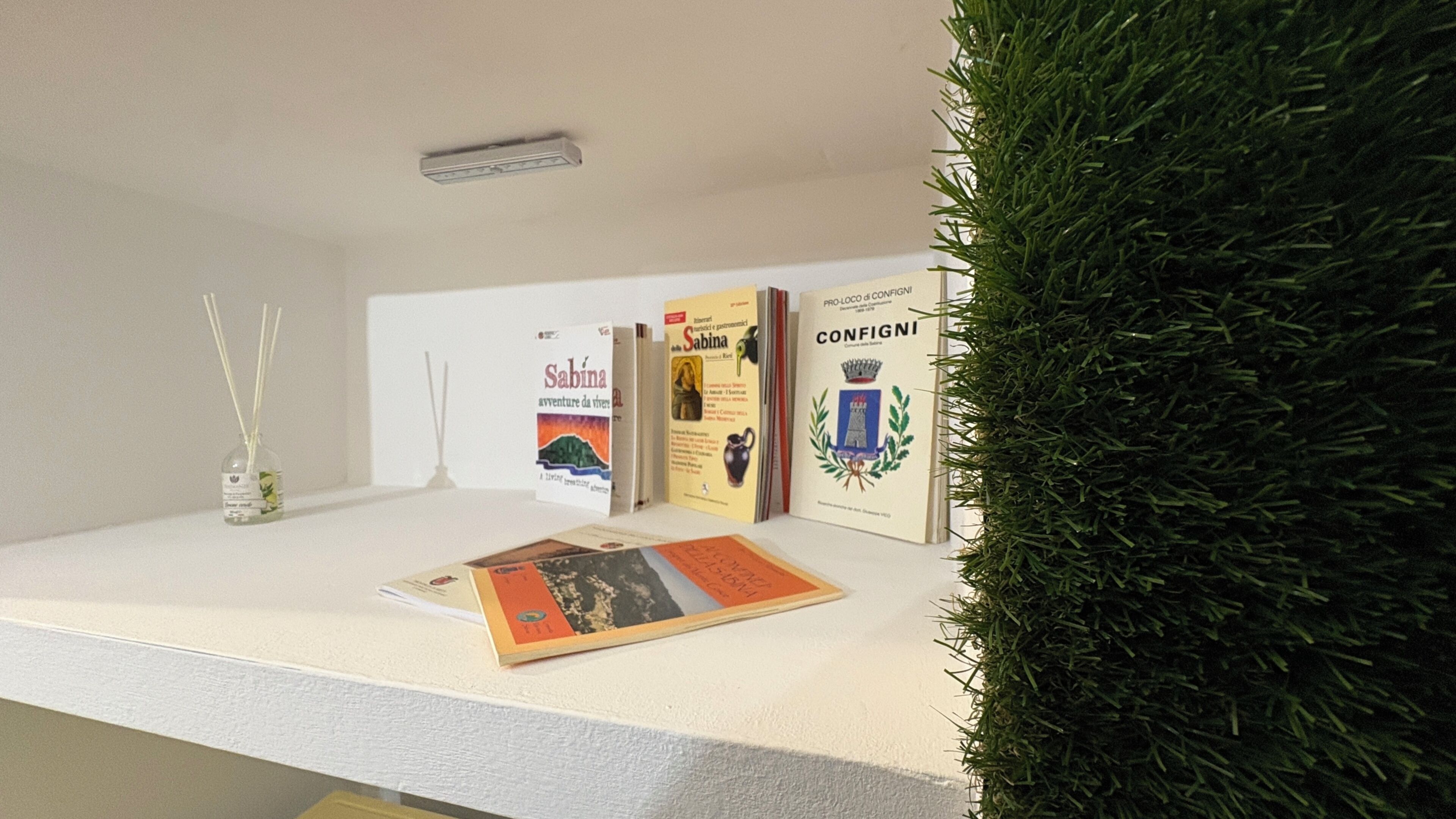 Een receptiebalie met een witte toonbank, een kleine plant en drie boeken met de titels 'Sabina', 'Configui' en 'Pro-Loco di Confinio'.