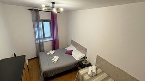 Uma cama de solteiro com almofadas e toalhas, uma mesa de cabeceira, uma janela com cortinas e um lustre no teto.