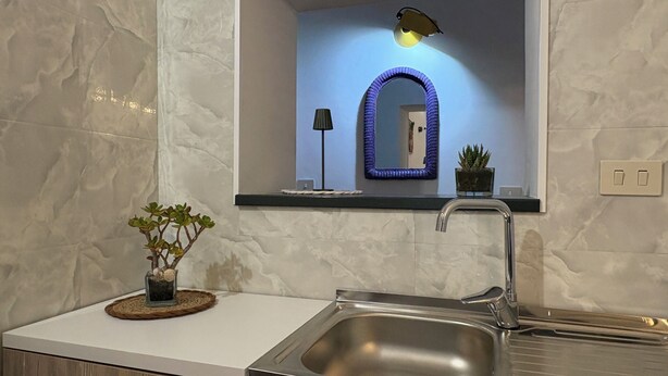 Cozinha moderna com pia de aço inoxidável, bancada preta, planta em vaso, espelho com moldura azul e luminária de parede.