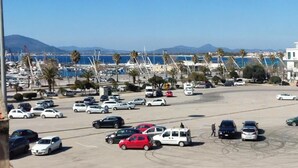A parking lot with cars, a marina, and mountains in the background. - algherohorizon (Alghero)