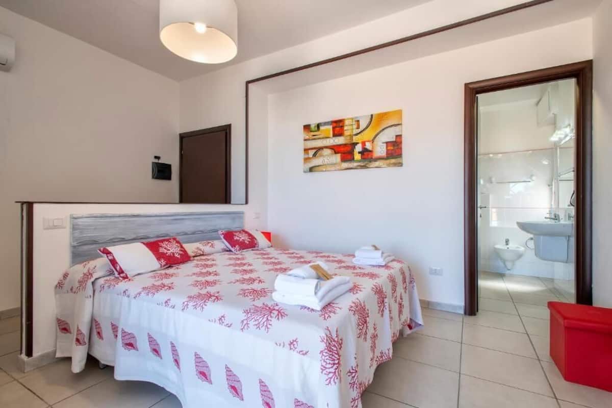 A bedroom with a bed, a painting, a bathroom visible through an open door, and a red bench.