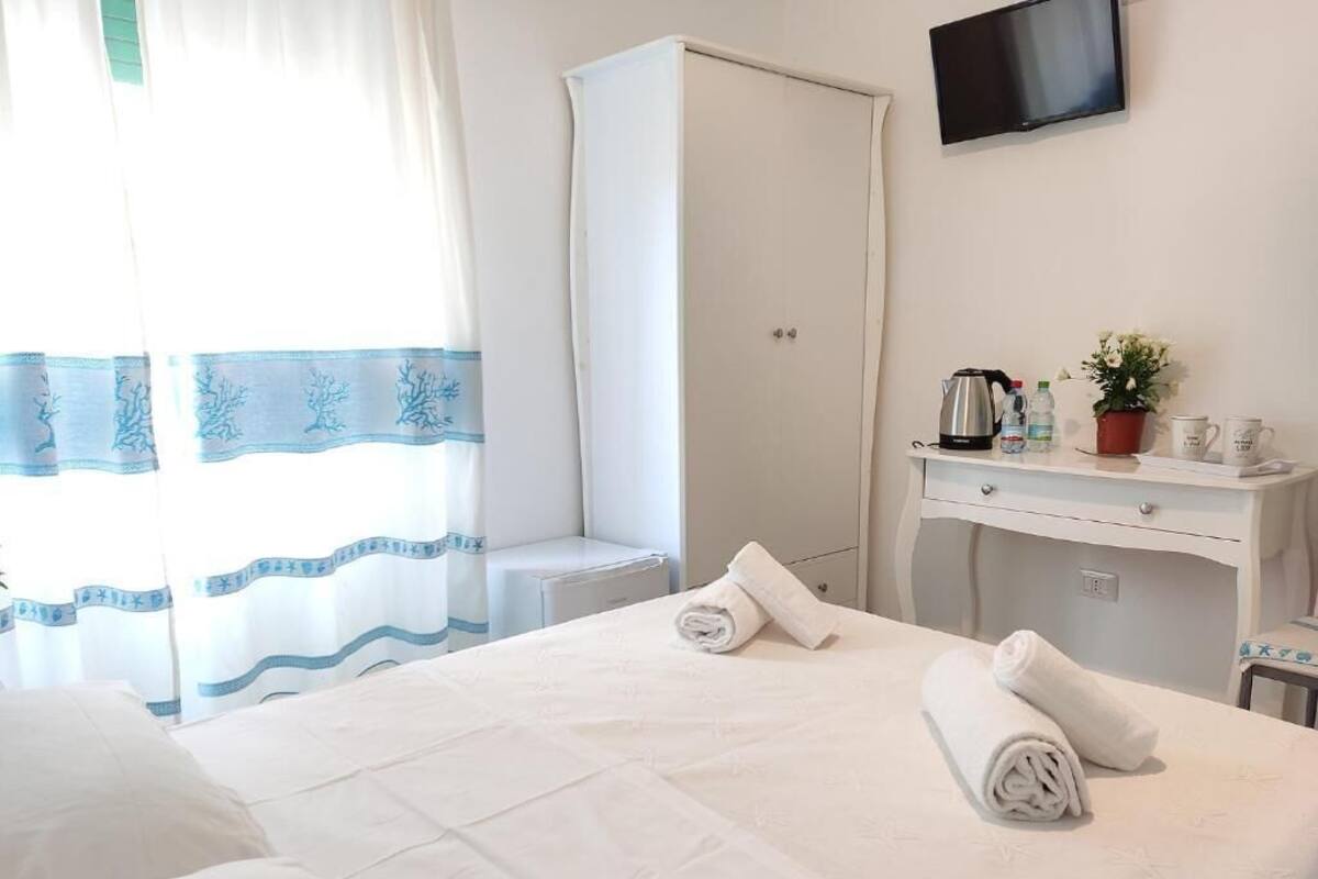A hotel room with a bed, a wardrobe, a desk with a flat-screen TV, a small table with a plant, and a window with curtains.