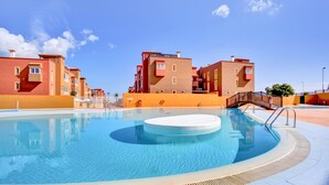 A swimming pool with a circular hot tub, surrounded by buildings with balconies and a clear sky. - Holiday Home 'Salitre y Lava' with Shared Pool and Private Terrace (Corralejo)