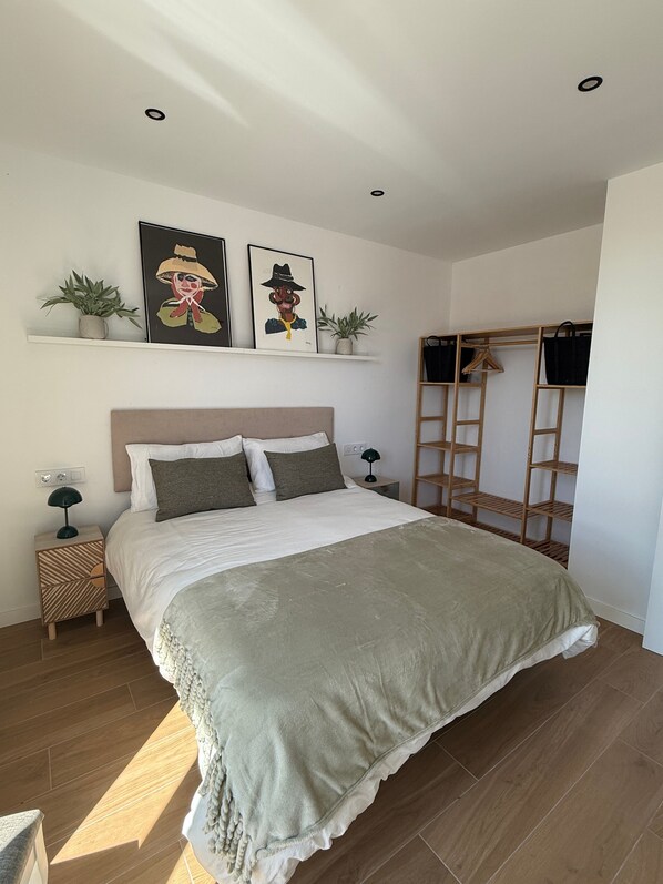 Un dormitorio con cama, mesita de noche, lámpara, estante con obras de arte, planta y armario de madera.