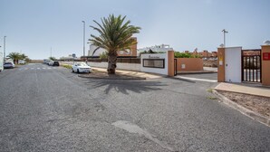 A street view with a gated entrance, palm trees, and parked cars. - Holiday Home 'Salitre y Lava' with Shared Pool and Private Terrace (Corralejo)