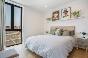 Een slaapkamer met een groot bed, een nachtkastje, een lamp, een raam met uitzicht op gebouwen en wanddecoratie.