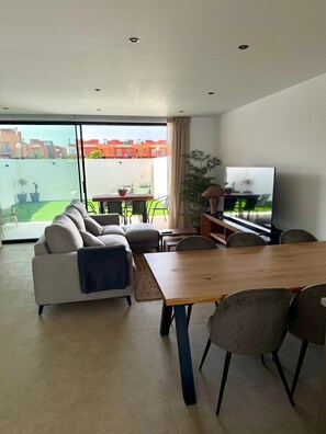 Una sala moderna con sofá, mesa de comedor, sillas, televisor y un balcón con vista a edificios.