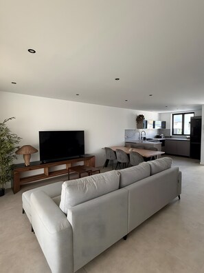 Una sala de estar moderna con un sofá grande de color gris, un televisor de pantalla plana, una mesa de centro de madera y un comedor con una cocina al fondo.