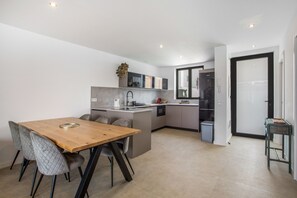 A modern kitchen with a dining area, featuring a wooden table and chairs, a built-in oven, and a small island with a sink. - Holiday Home 'Salitre y Lava' with Shared Pool and Private Terrace (Corralejo)