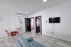 Una sala de estar moderna con un sofá, una mesa de comedor con sillas, un televisor montado en la pared y un equipo de aire acondicionado.