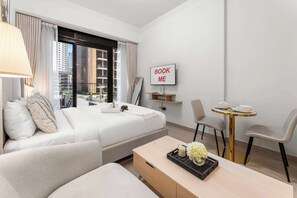 A modern hotel room with a bed, a small dining table with chairs, a TV mounted on the wall, and a window with curtains. - Free housekeeping visits during your stay - Peaceful Studio for 2 in Meydan! (Meydan)