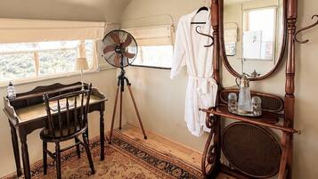 A room with a wooden desk, chair, and a large mirror with a stand. There's a fan, a small table with a lamp, and a patterned rug.