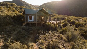 A wooden cabin with a deck, surrounded by dense shrubbery and hills.