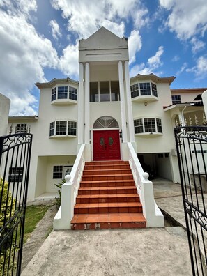Un immeuble à plusieurs étages, avec une porte rouge au centre, une façade blanche et un escalier menant à l’entrée.