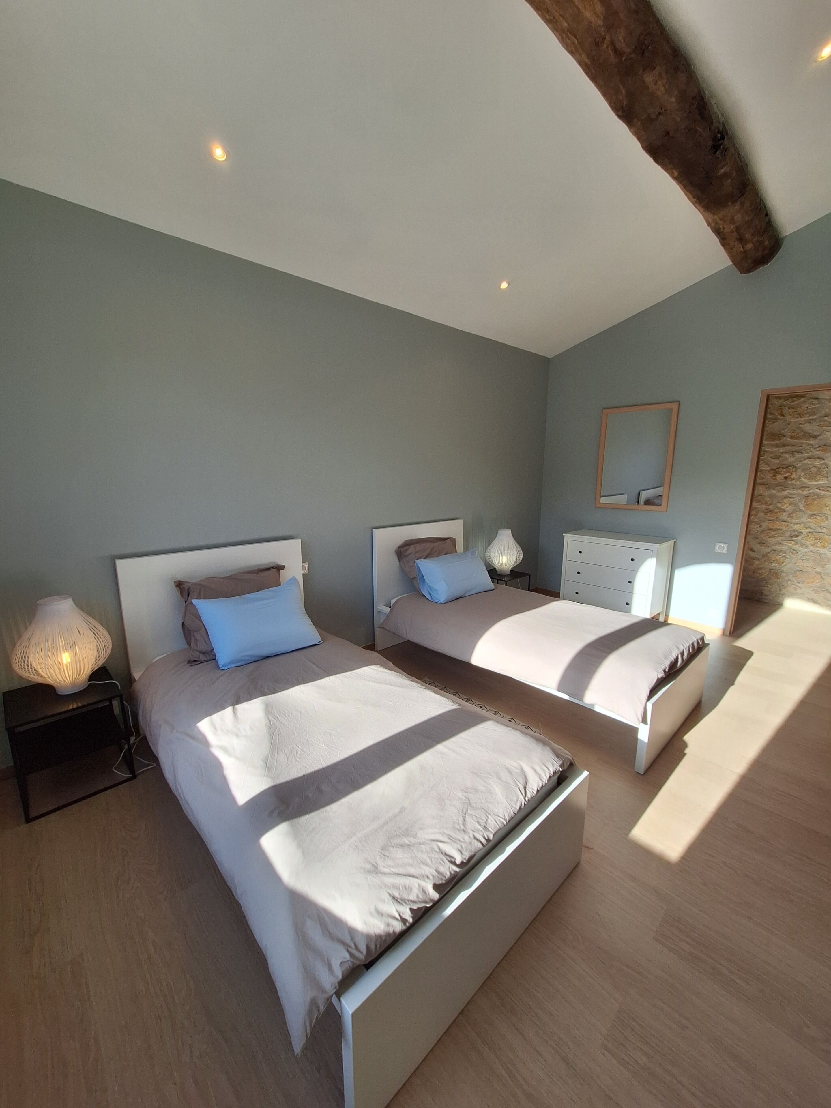Un dormitorio con dos camas, una mesita de noche con lámpara, un aparador, un espejo y una pared de piedra.